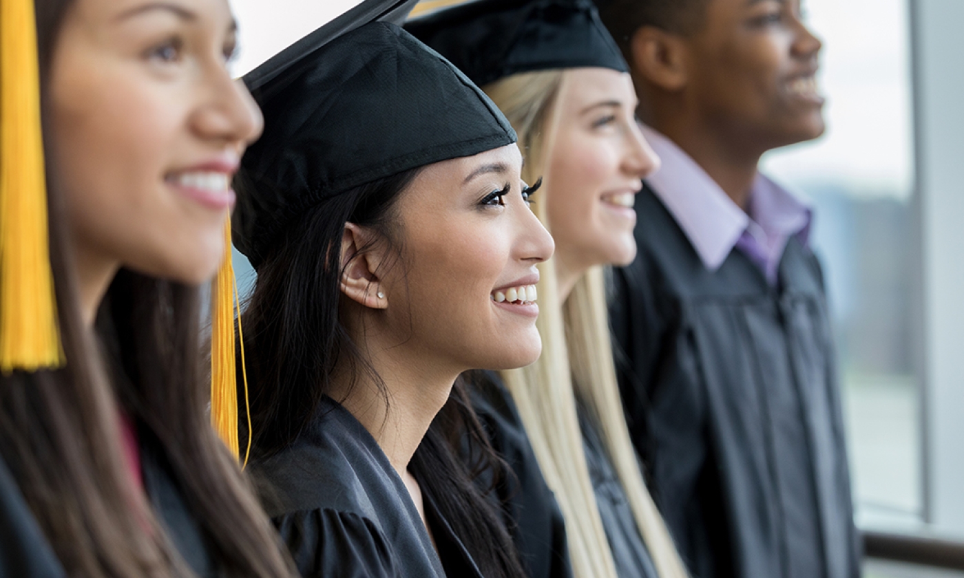 hree high school girls and one boy standing in a row wearing a cap and gown for  graduation, smiling. 