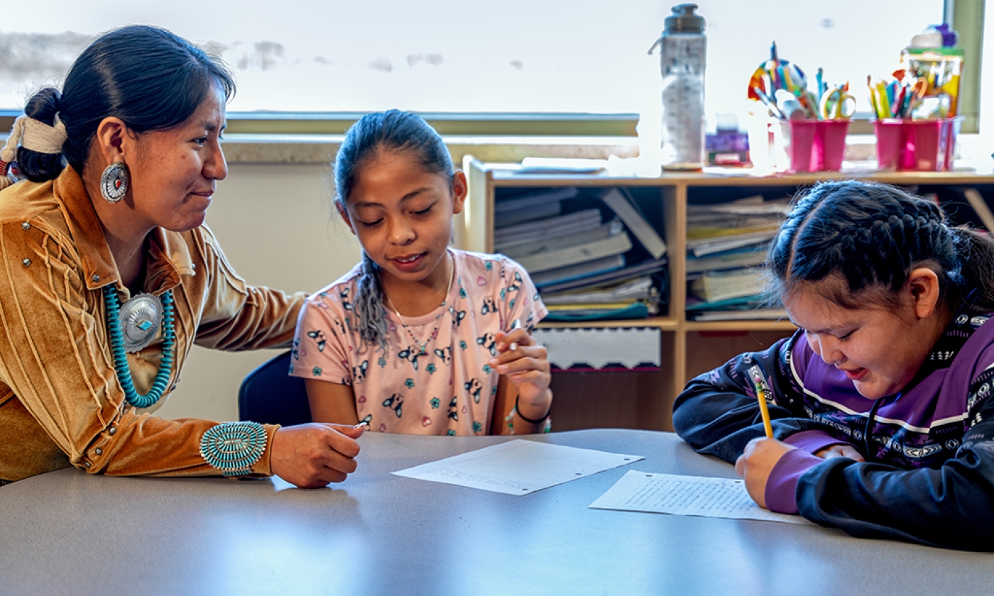 An indigenous woman assisting elementary age students in a classroom as they sit at a table together.